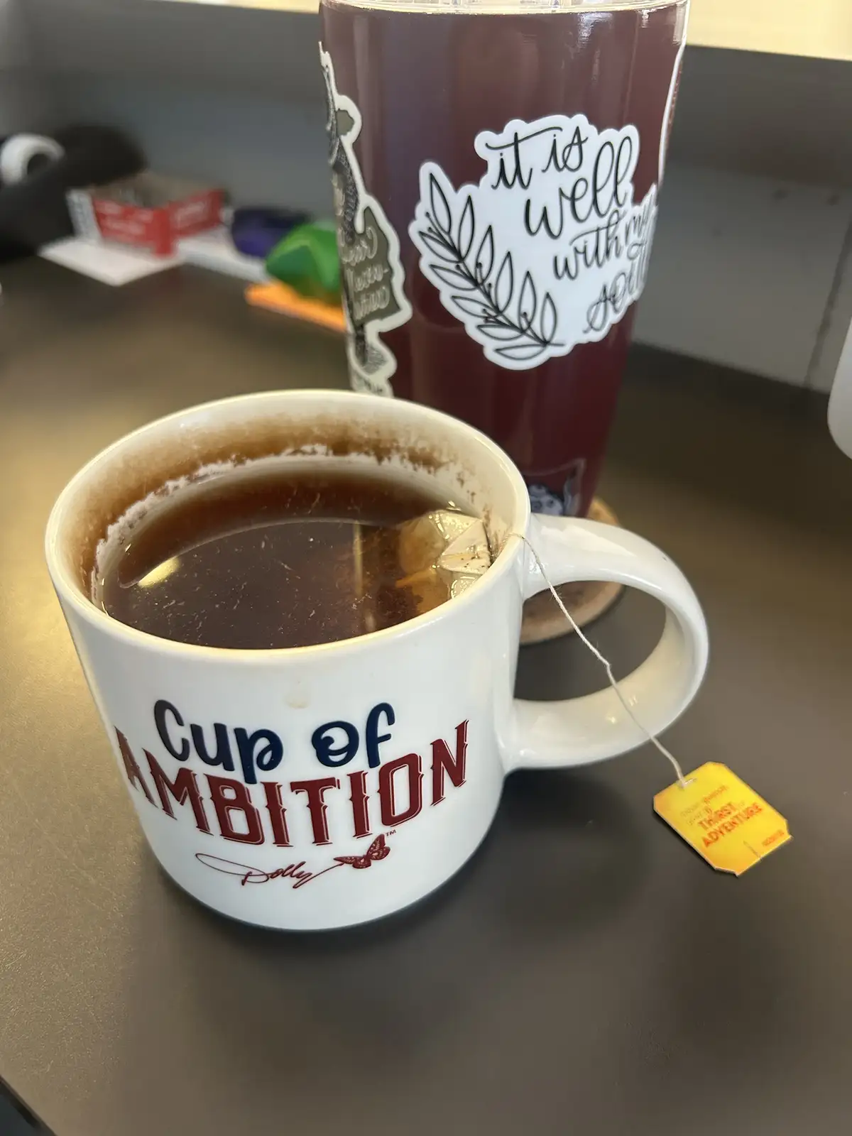 A white ceramic mug with the text 'Cup of AMBITION' and a butterfly logo sits on a desk, filled with tea and a steeping tea…
