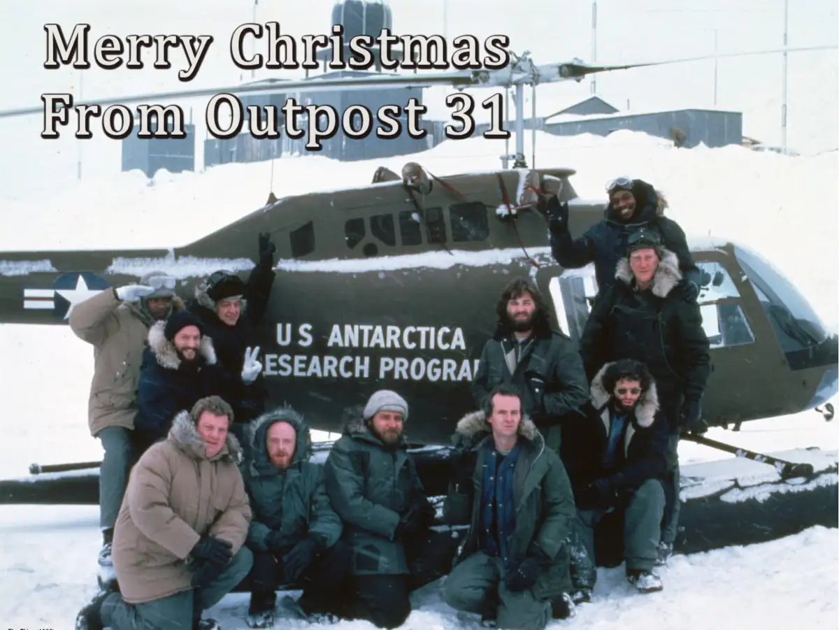 A group of men in heavy winter gear pose cheerfully in front of a U.S. Antarctica Research Program helicopter in a snowy…