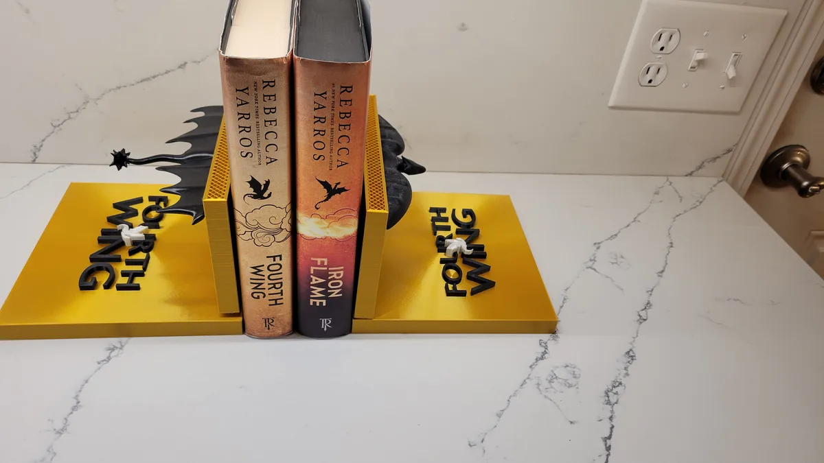 Two books by Rebecca Yarros, 'Fourth Wing' and 'Iron Flame', are held upright by custom yellow bookends shaped like dragon…