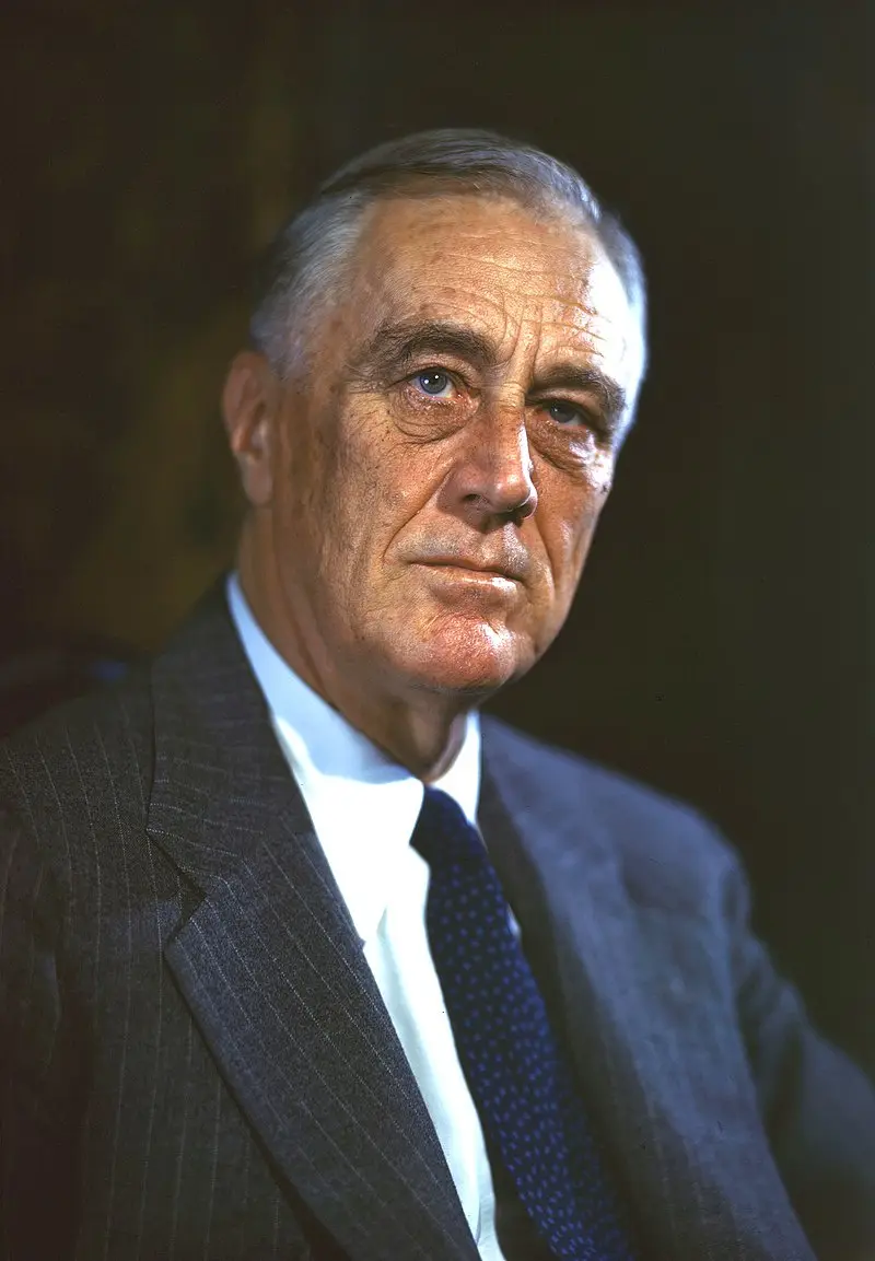 a formal portrait of an older man with gray hair, wearing a dark pinstripe suit, white shirt, and blue patterned tie…