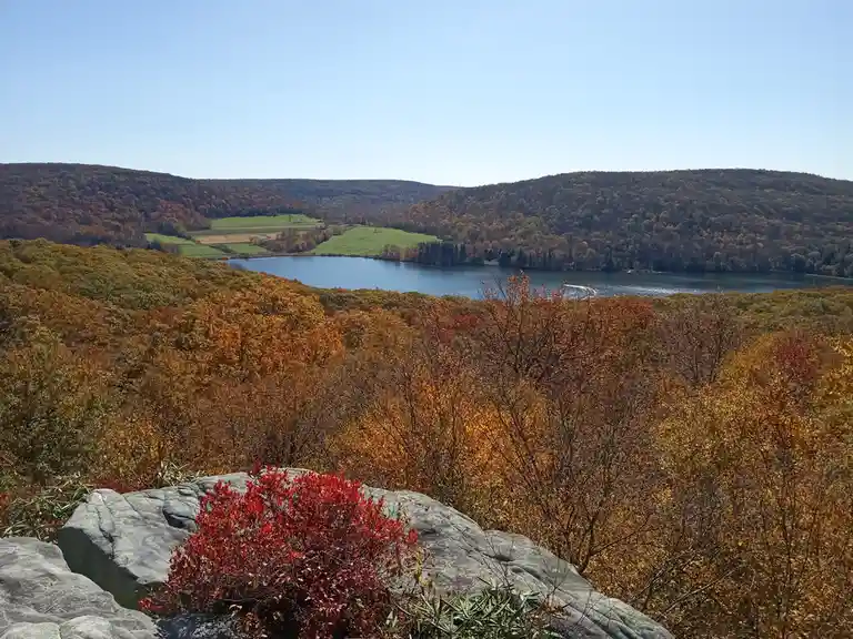 A scenic autumn landscape with a lake surrounded by hills covered in orange, red, and yellow foliage under a clear blue sky…