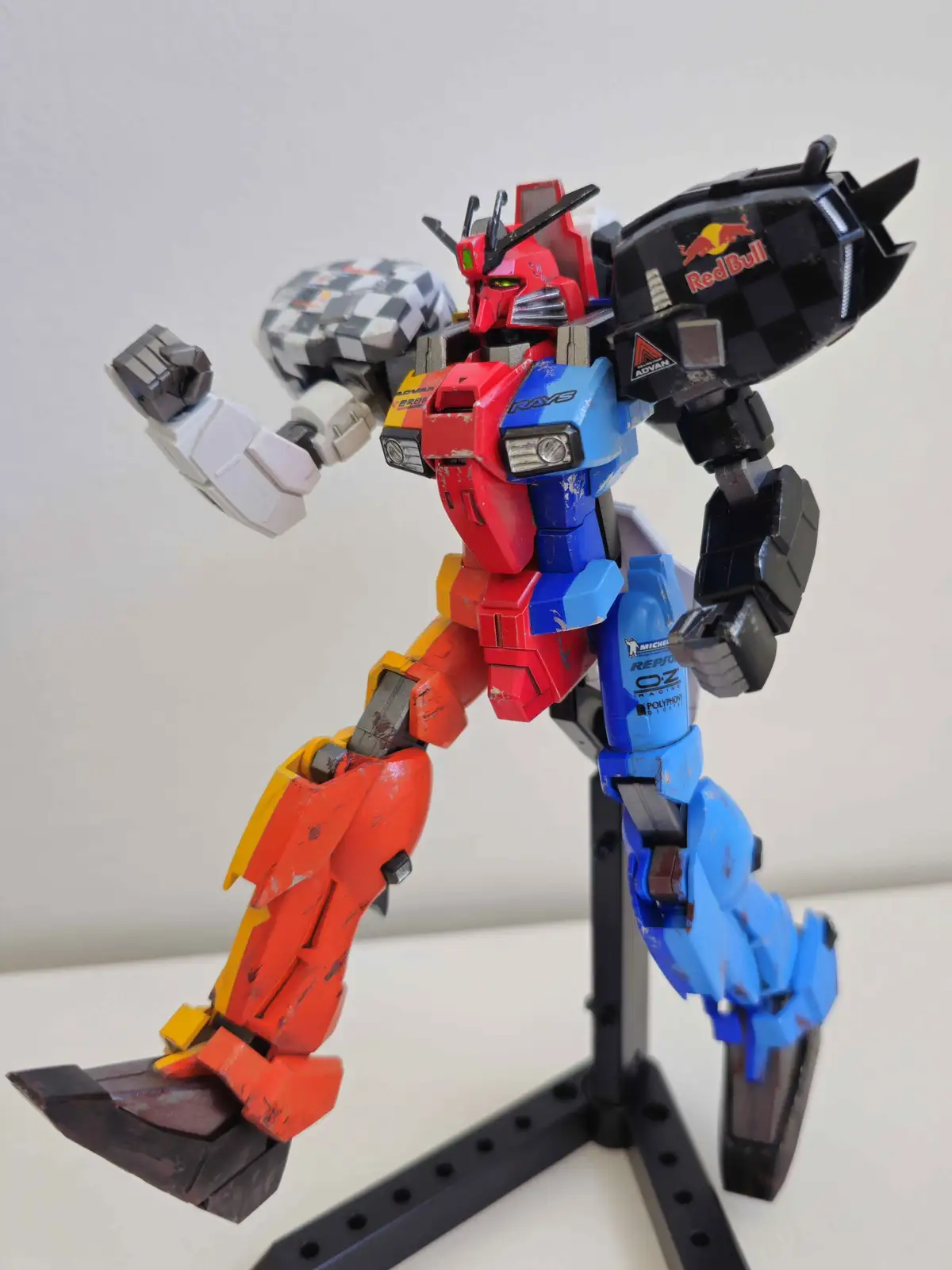 A red, blue, and orange robot model, resembling a Gundam, stands on a black display stand with a dynamic pose. Its right arm…