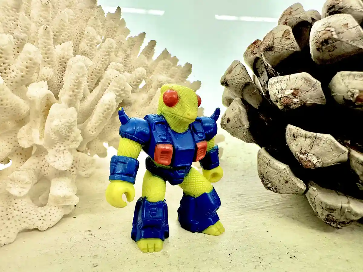 A small, yellow and blue plastic action figure with red eyes stands between a white coral-like structure on the left and a…