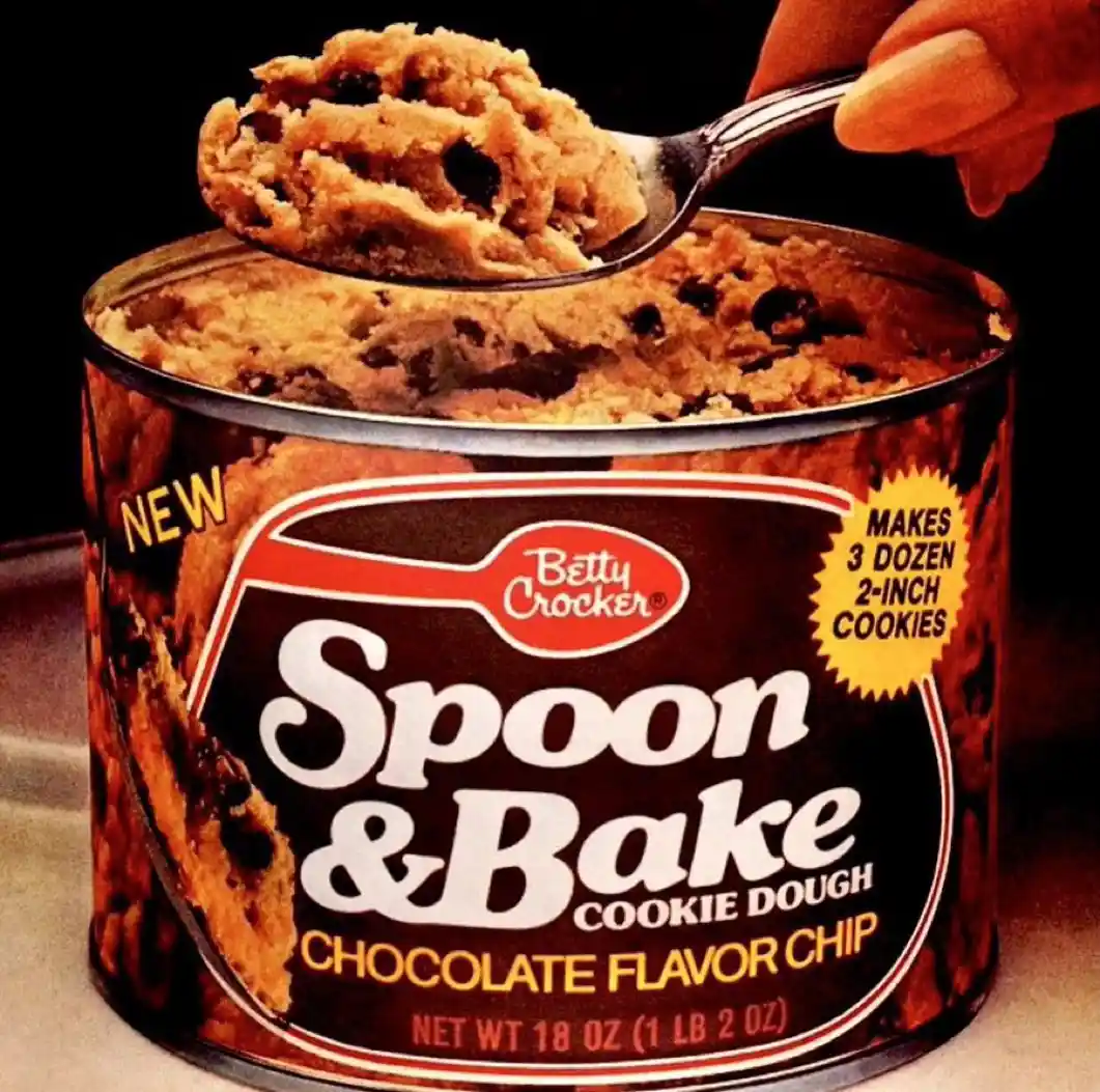 A vintage-style advertisement image shows a hand holding a spoon lifting a scoop of chocolate chip cookie dough from a can…