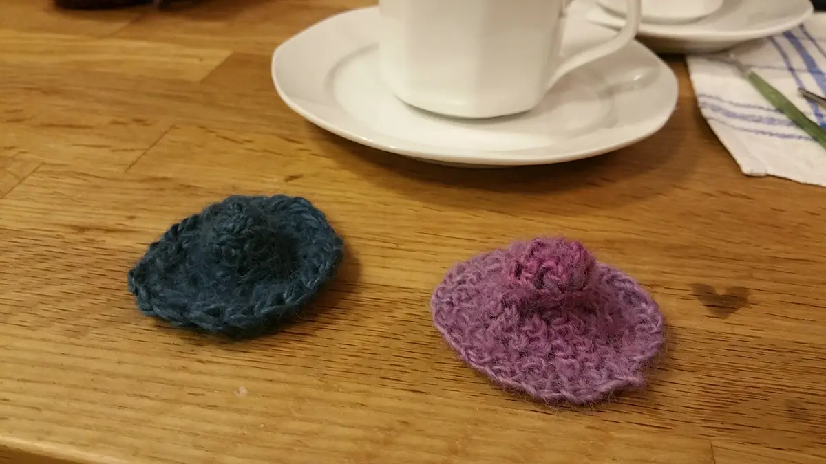 Two small, crocheted, hat-like items—one blue and one purple—rest on a wooden table, with a white coffee cup and saucer in…