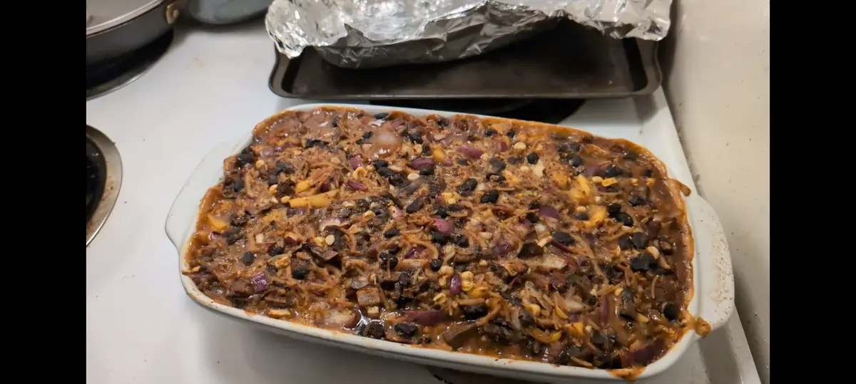 A white ceramic baking dish sits on a white stovetop, filled with a messy, uneven casserole topped with black beans, red…
