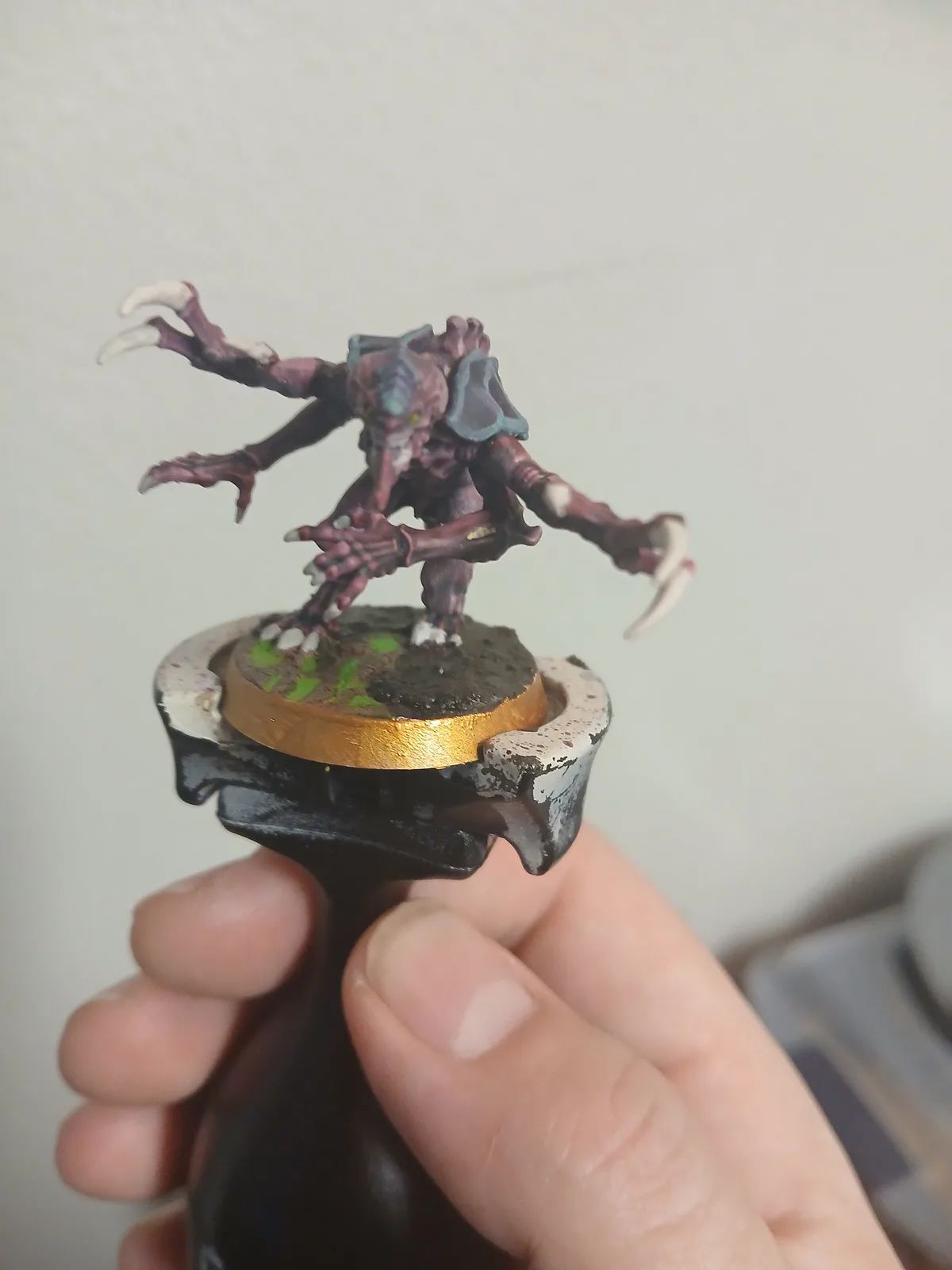 A hand holds a painted miniature figurine of a grotesque, multi-limbed creature with claws and green eyes, mounted on a…