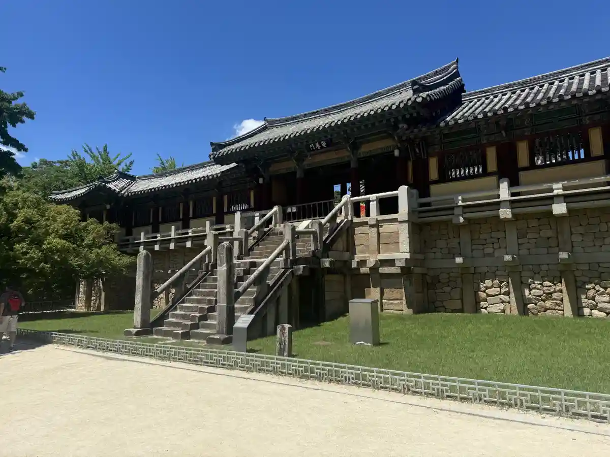 A bright, sunny photo of a traditional Korean temple gate with a stone staircase leading up to it, surrounded by green grass…