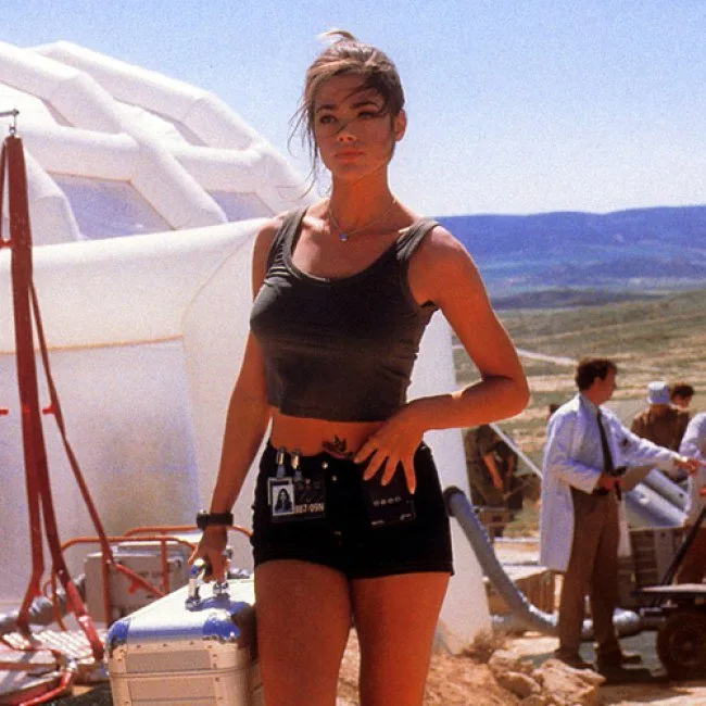 A woman in a black crop top and shorts stands confidently outdoors, holding a suitcase, with a white dome structure and…