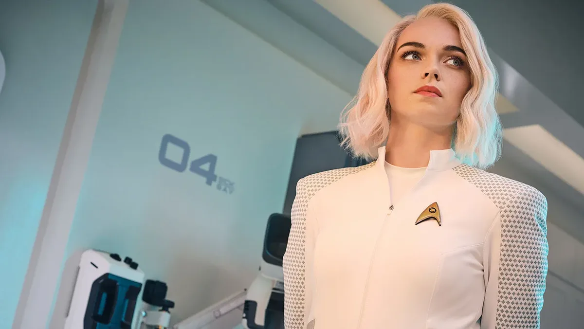 a woman with short blonde hair wearing a white Starfleet uniform with a gold delta-shaped insignia looks upward with a…