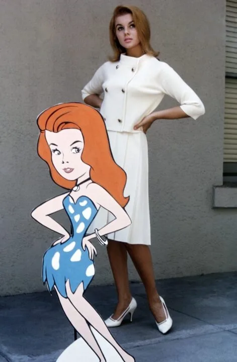 A real woman in a white dress and heels stands next to a cardboard cutout of a cartoon character, both striking similar…