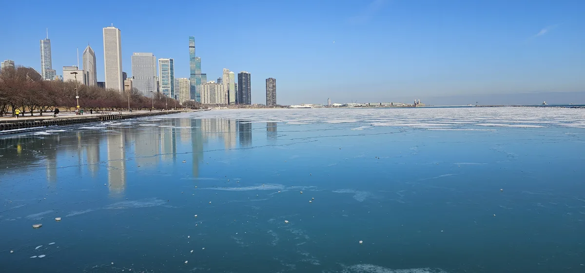 A wide shot of the Chicago skyline with its skyscrapers reflected in a partially frozen lake under a clear blue sky. People…