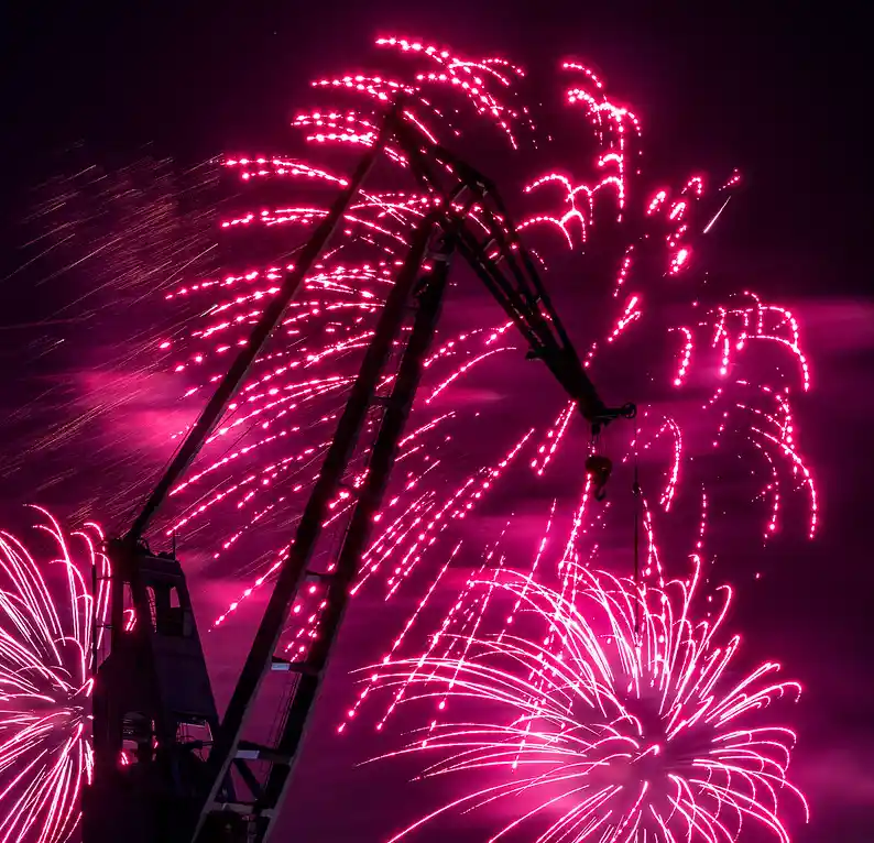 A dark silhouette of a large industrial crane stands against a night sky filled with vibrant pink and magenta fireworks…