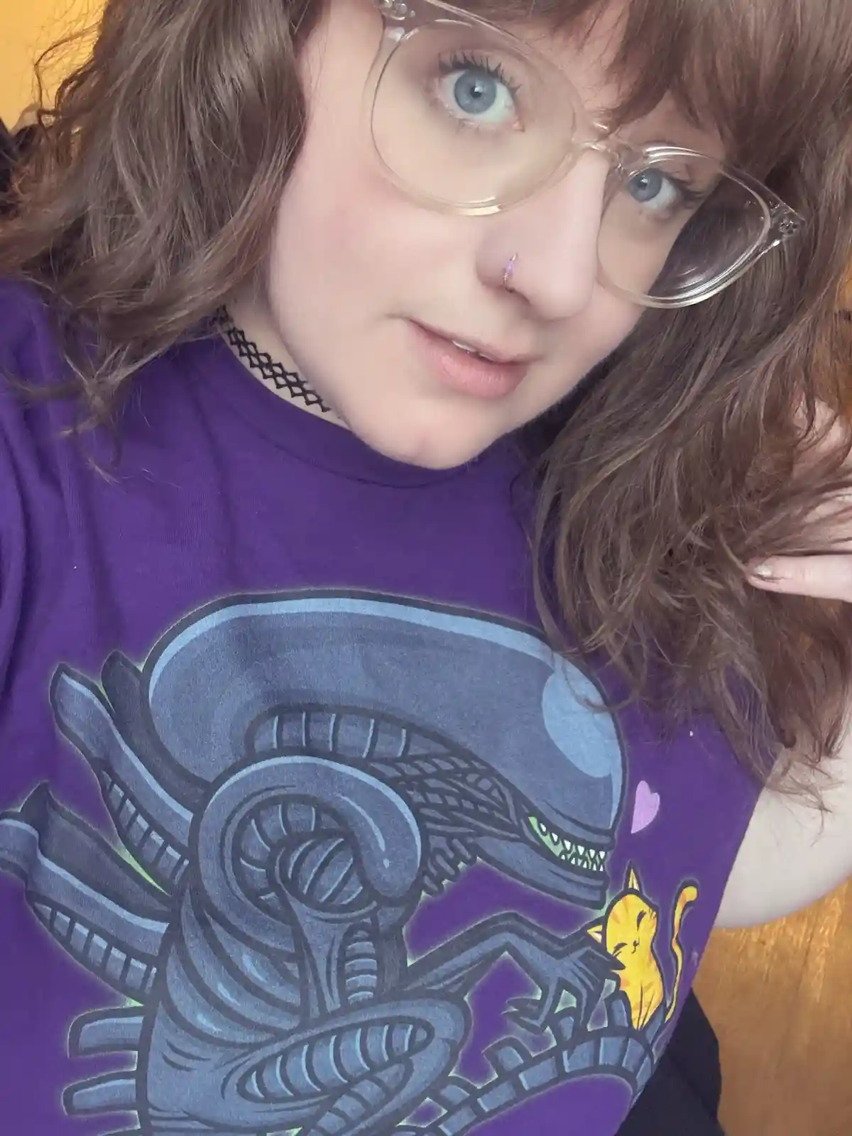A person with brown hair, clear-framed glasses, and a nose ring looks at the camera while wearing a purple t-shirt. The…