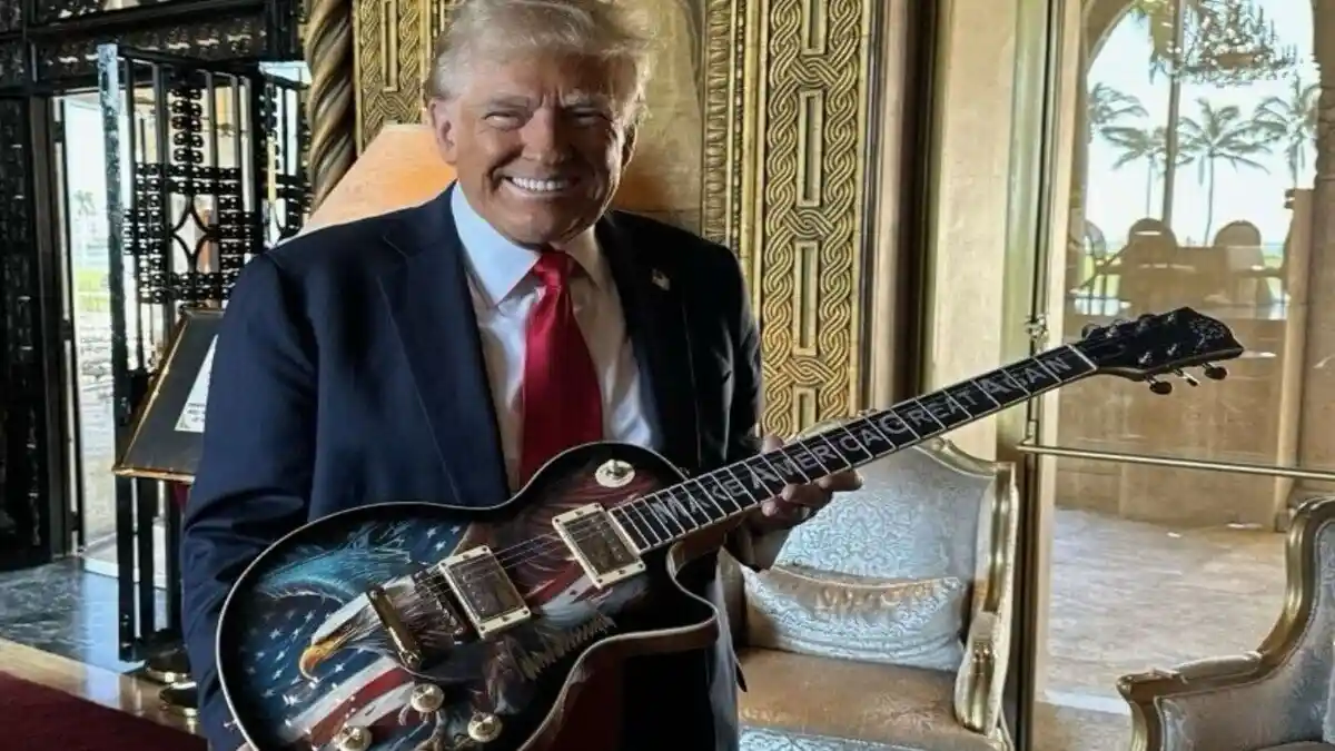 a man in a dark suit and red tie smiles while holding an electric guitar with 'make america great again' written along the…