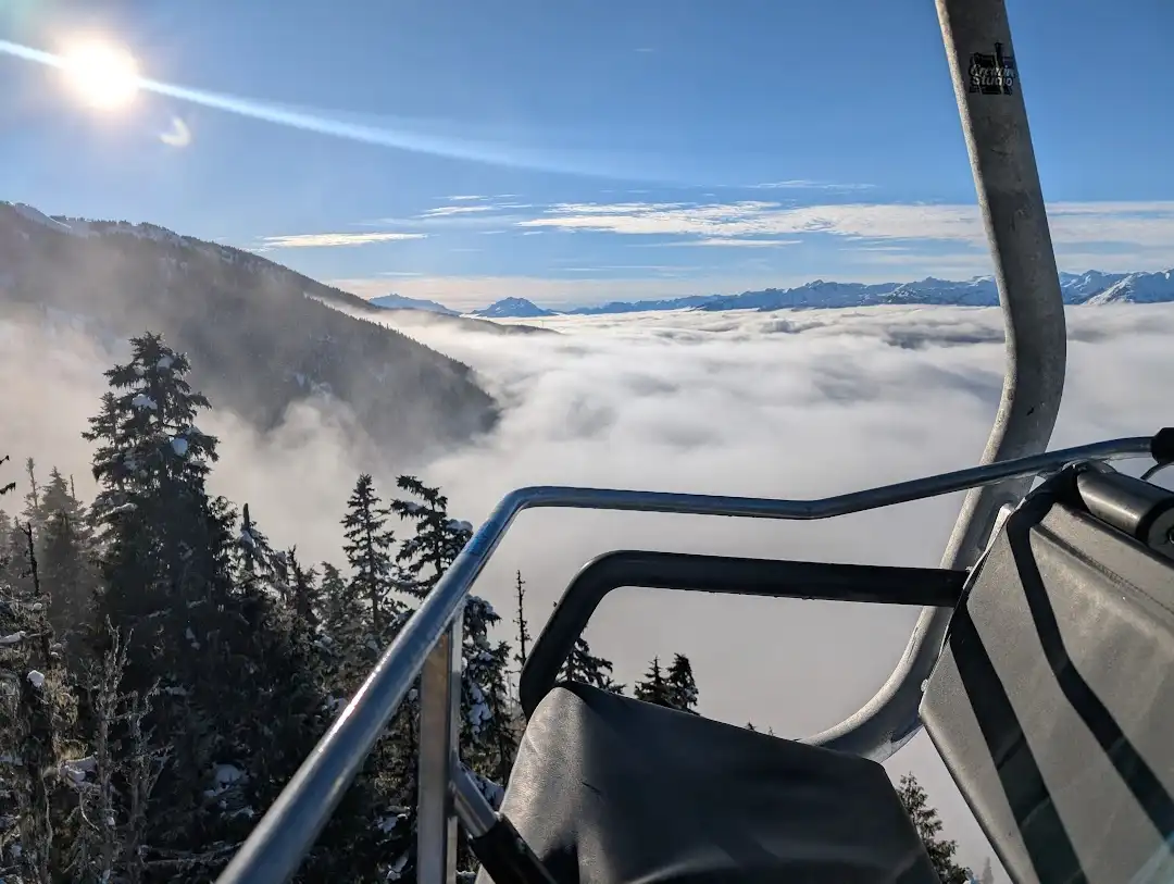 A first-person view from a ski lift chair, looking out over a vast sea of clouds with snow-covered mountains and evergreen…