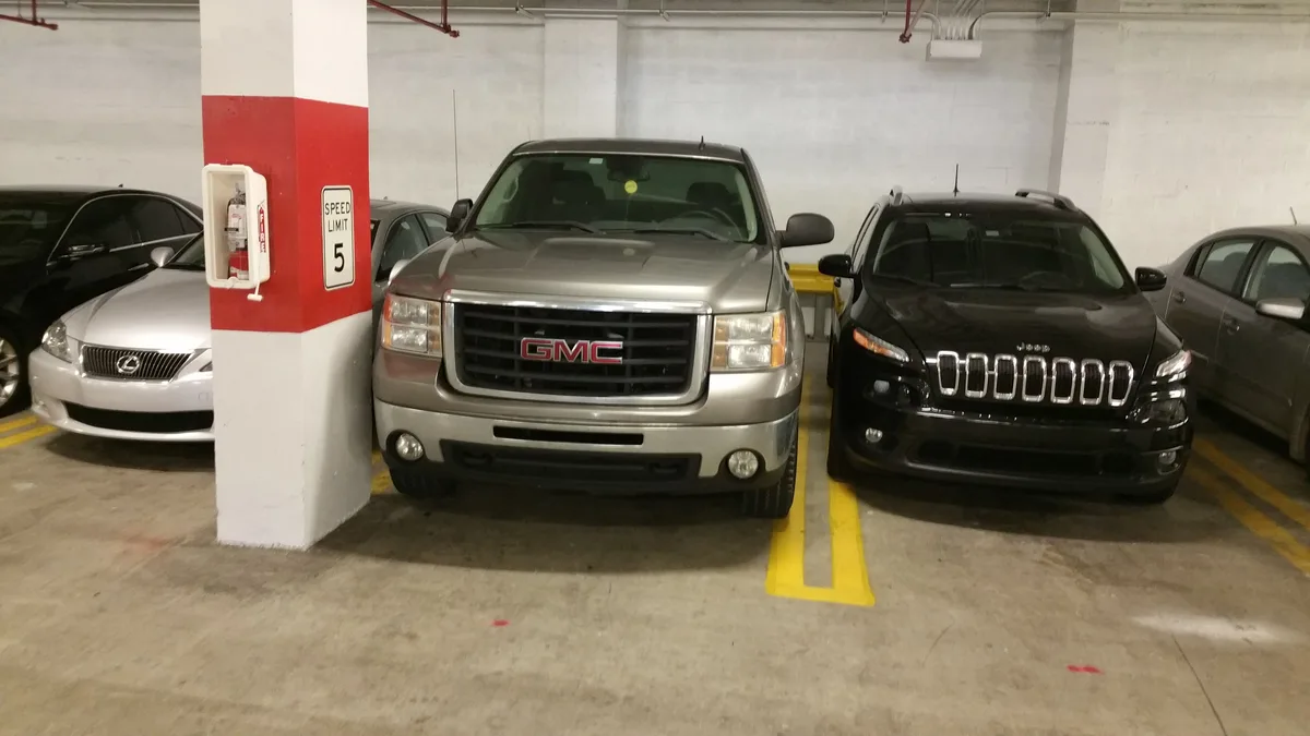 A silver GMC truck is parked in an underground garage next to a black Jeep, with a red-and-white pillar beside it displaying…