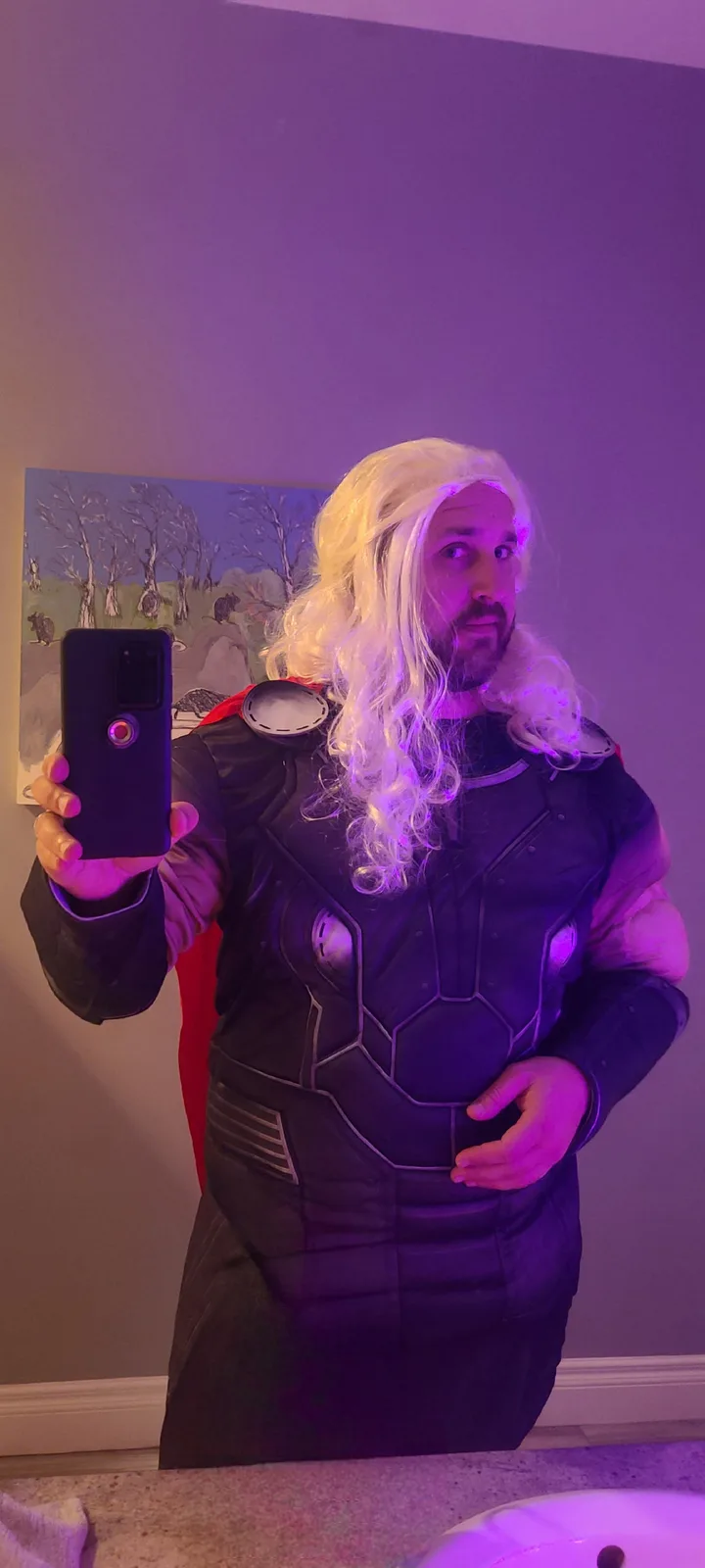 A man in a Thor costume with a long blonde wig and black armor takes a mirror selfie in a purple-lit room, looking confused…