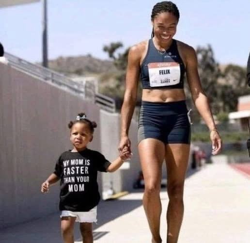 A tall, smiling adult woman in athletic gear with a 'FELIX' race bib walks hand-in-hand with a small child wearing a black…