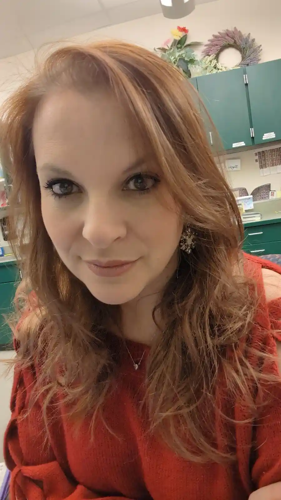 A woman with long, wavy reddish-brown hair and a subtle smile looks directly at the camera. She wears a red off-the-shoulder…