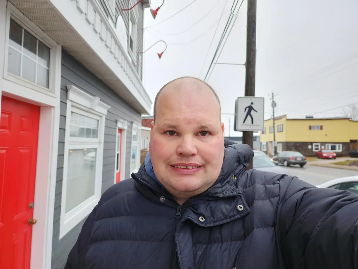 A man with a shaved head wearing a dark puffer jacket takes a selfie outdoors on a street with buildings and cars in the…