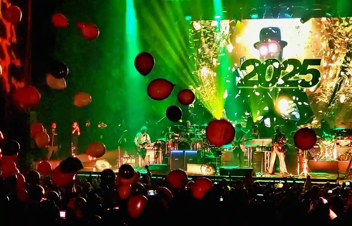 A concert stage bathed in green light with balloons floating in the air, featuring a large screen displaying the year '2025'…