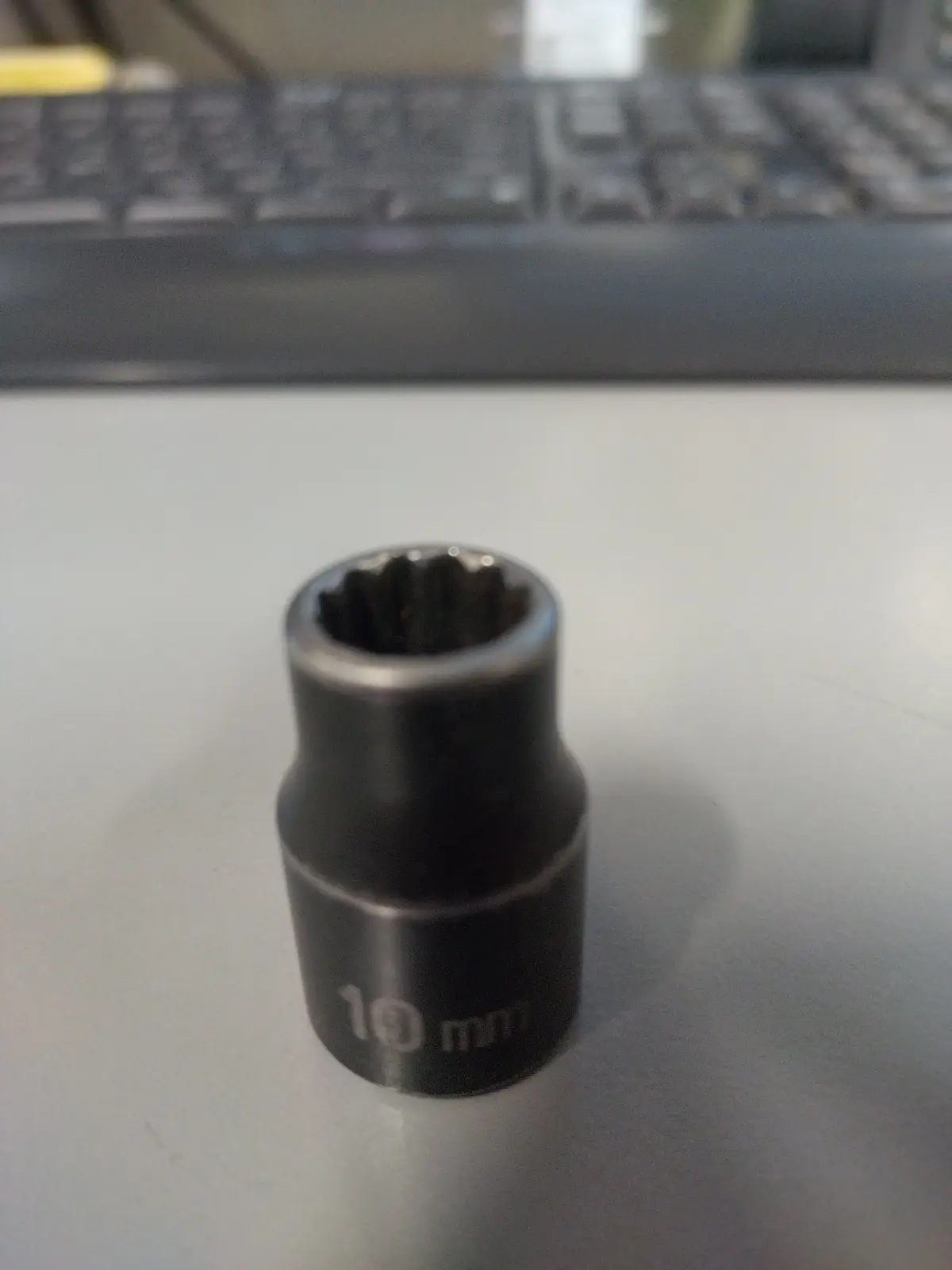 A close-up photo shows a black 10 mm socket wrench bit sitting on a white desk surface, with a blurred computer keyboard…
