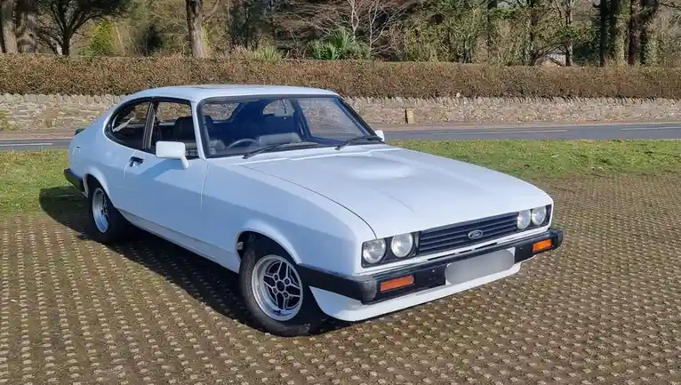 A light blue classic Ford Capri is parked on a grassy area with a patterned ground cover, facing the viewer. The car has a…