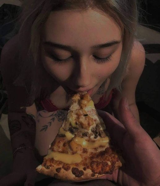 A person with light-colored hair and visible tattoos on their arm is leaning in to eat a large slice of cheese-heavy pizza…