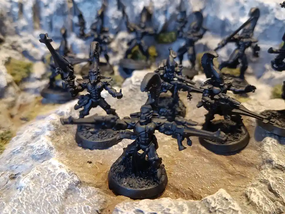 A close-up photo shows a group of unpainted black miniature figurines, likely from a tabletop wargame, standing on rocky…