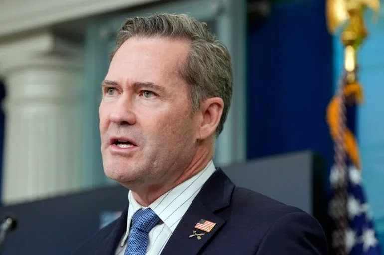 a man in a dark suit and blue tie with an american flag pin on his lapel stands in front of a blurred background with an…