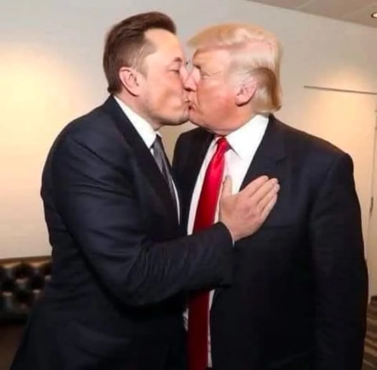 Two men in suits are kissing each other on the cheek in a close-up photo. The man on the left, with dark hair, is wearing a…