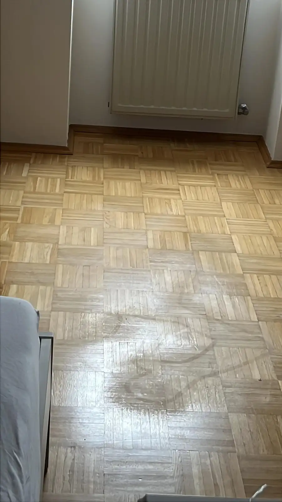 A photo of a wooden parquet floor with a large, shiny, wet-looking stain in the center, likely from a spill. The floor is…