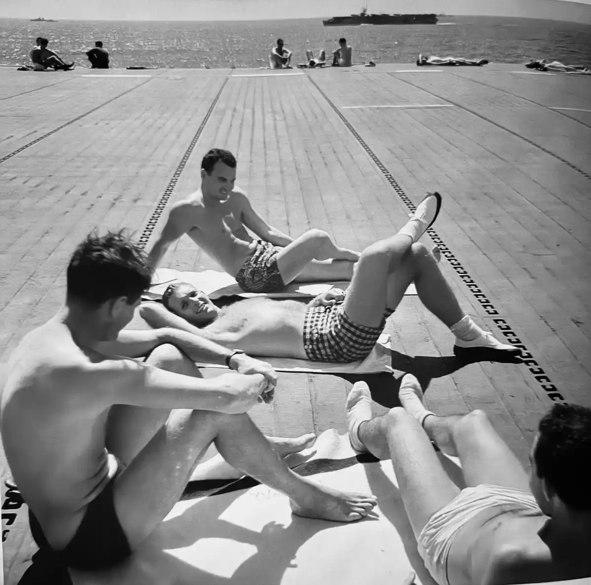 A black-and-white photo shows several shirtless men relaxing on a wooden flight deck, likely an aircraft carrier, with the…