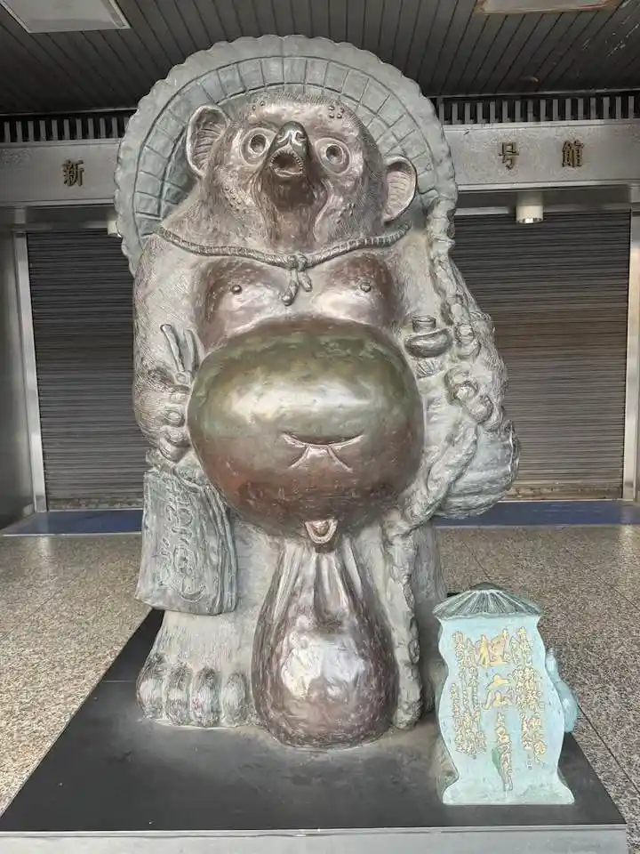 一座青铜熊雕像，体型肥硕，头顶光环，双手捧着一个巨大的球形物体，球体下方有类似尿道的开口，雕像旁立着一块刻有日文的石碑。背景是新宿站的卷帘门和标有“新宿駅”字样的建筑结构。雕像表情夸张，整体造型滑稽，常被网友用于调侃或表达“憋不住了”的状态。