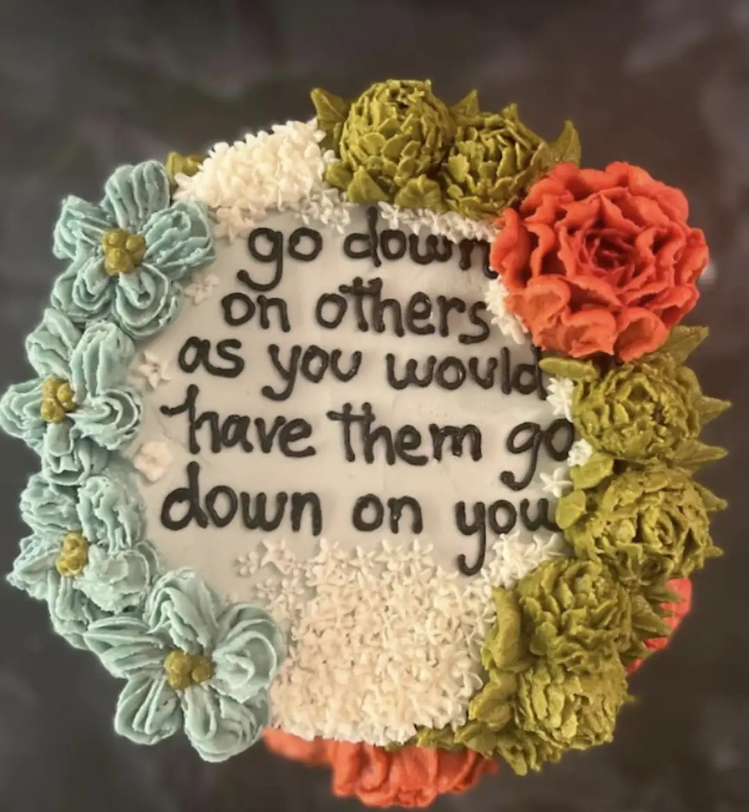 A round cake with white frosting is decorated with piped flowers in teal, white, olive green, and coral red. The center of…