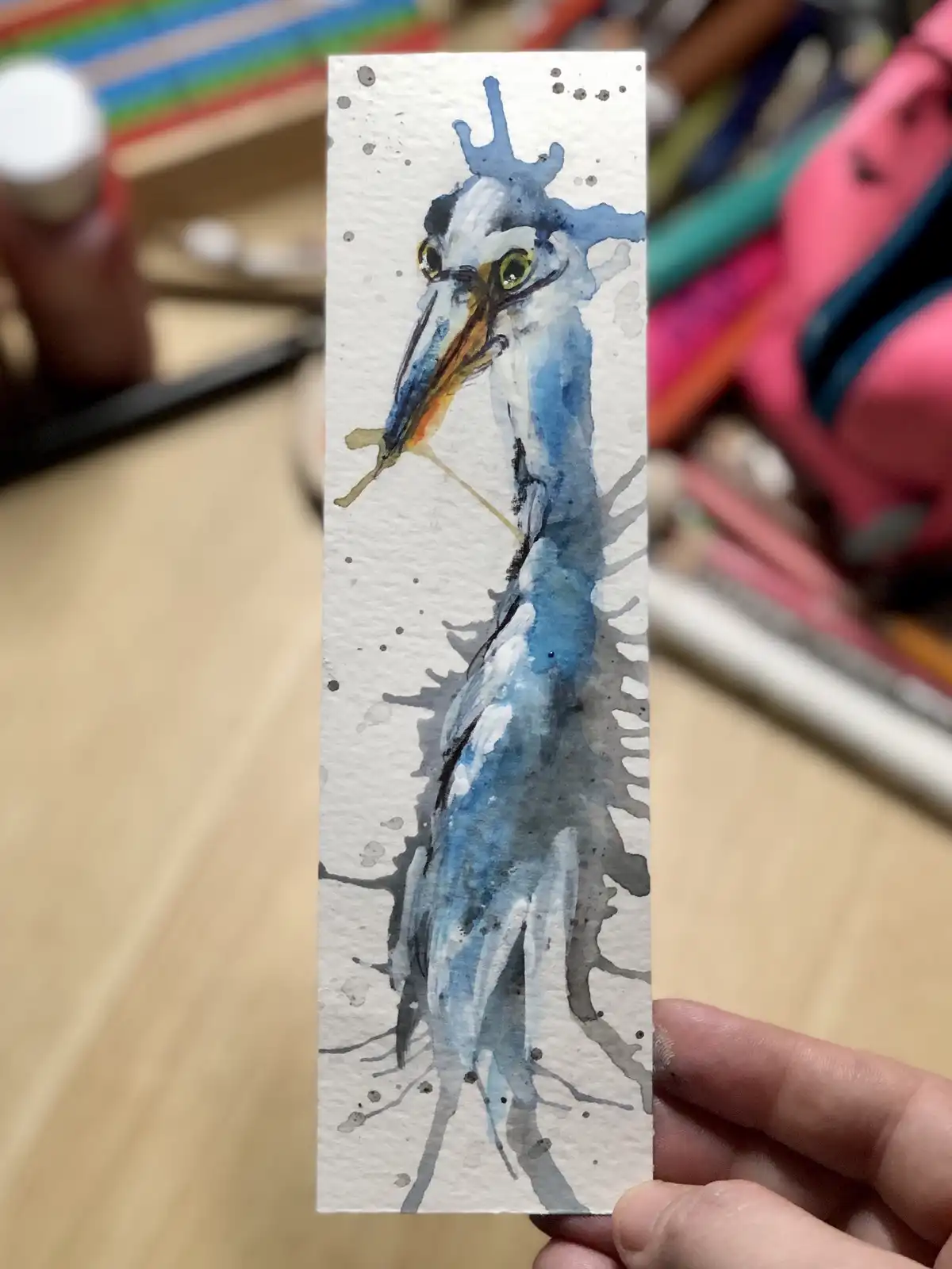 A hand holds up a small watercolor painting of a heron, rendered in blue, white, and gray with splatters and a crown-like…