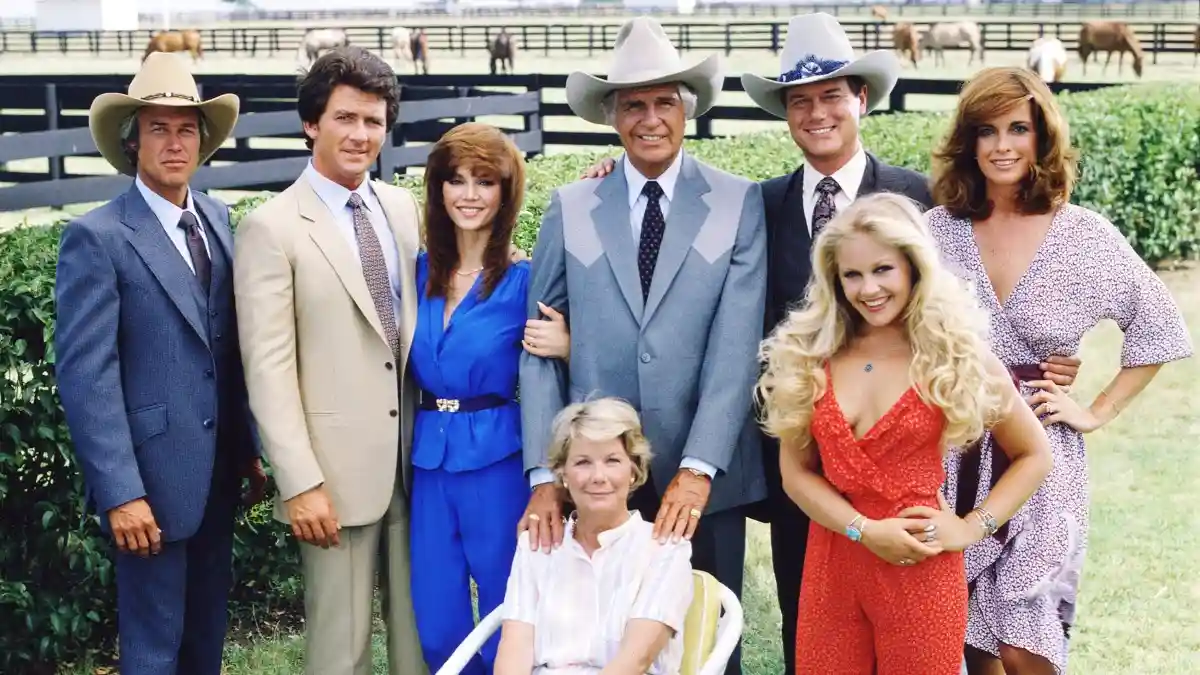 a promotional photo of the cast of 'The Dukes of Hazzard' standing outdoors in front of a horse pasture, dressed in…