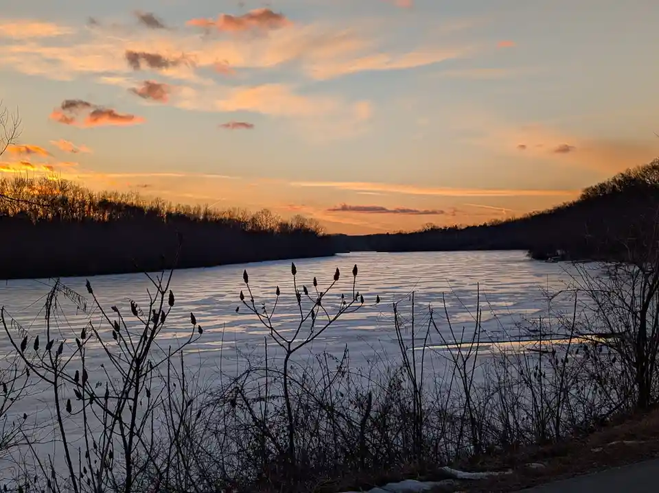 A serene winter sunset over a frozen lake, with bare branches in the foreground silhouetted against a sky painted with…