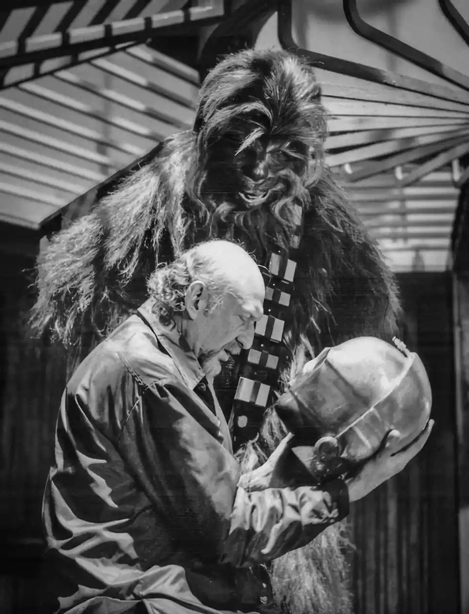 A black-and-white photo shows actor Peter Mayhew in the Chewbacca costume, holding a helmet while an older man with curly…