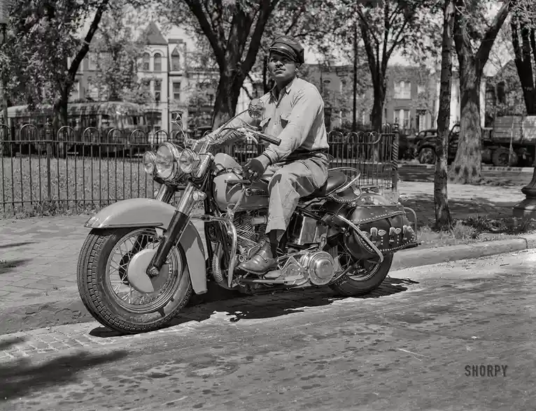 A black-and-white historical photo shows a man in a uniform and cap sitting on a vintage motorcycle parked on a paved…