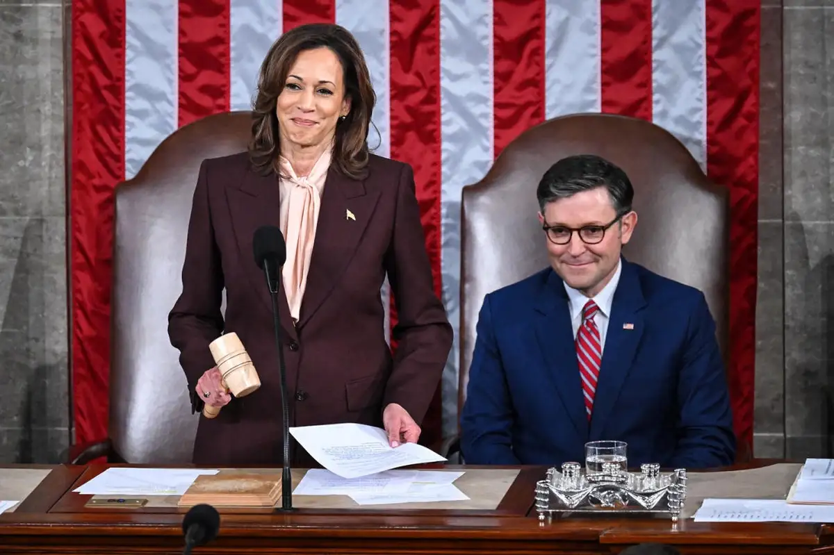 A photo of Kamala Harris standing at a podium holding a gavel and papers, smiling subtly, with Mike Johnson seated beside…