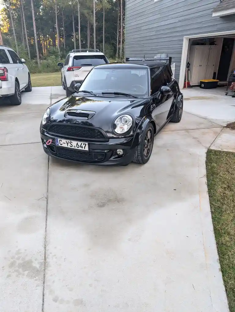 A black modified Mini Cooper is parked on a concrete driveway in front of a gray house with an open garage. Two white SUVs…