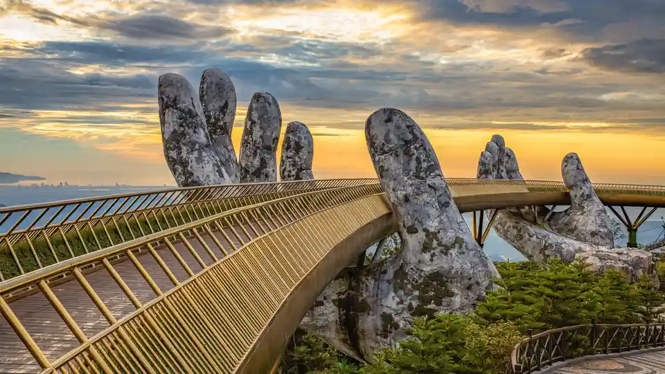 A golden pedestrian bridge with a lattice railing is supported by two massive, weathered stone hands emerging from the…