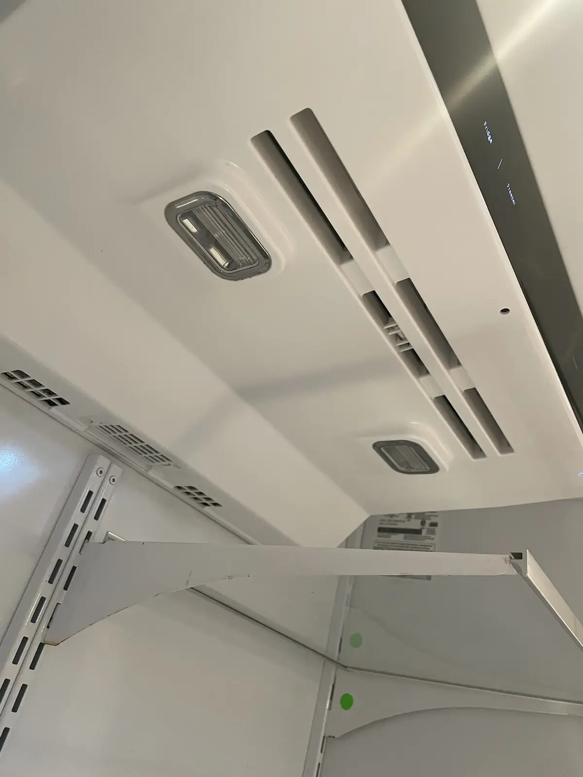 A low-angle photo looking up inside an empty, white refrigerator with visible vents, lights, and a digital display showing…