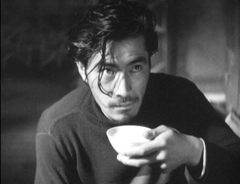 A black-and-white photo shows a man with dark, messy hair holding a small white bowl, looking intensely at the camera. His…