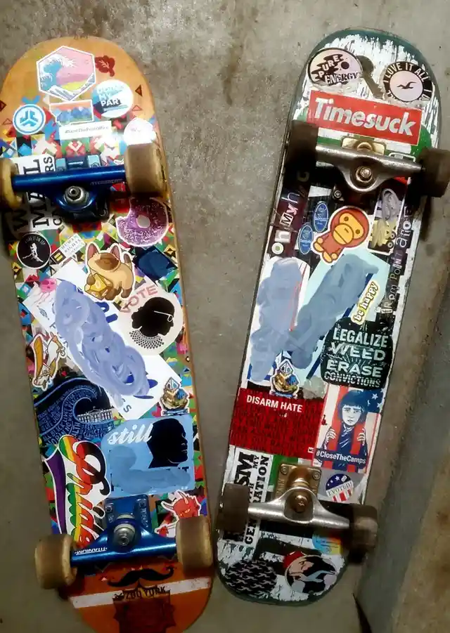 Two skateboards lie side by side on a concrete floor, both heavily covered in political and activist stickers. The left…