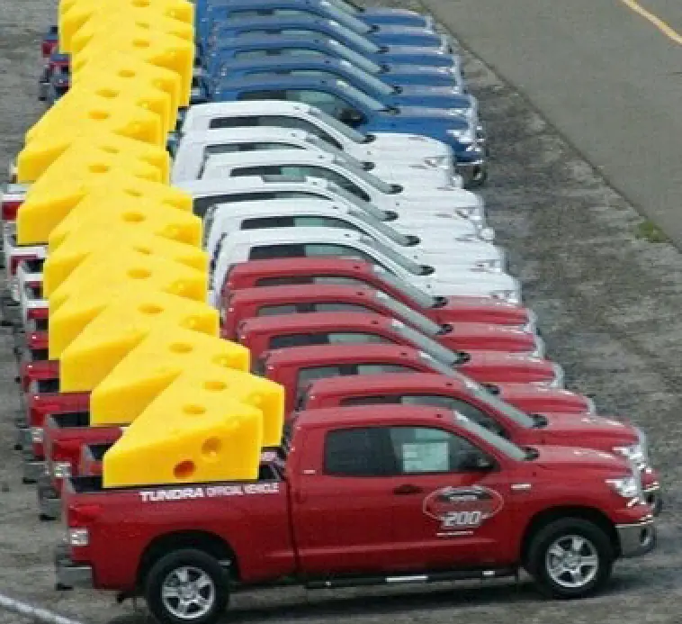 Rows of red, white, and blue pickup trucks parked in a line, each loaded with a giant yellow wedge of Swiss cheese in the…