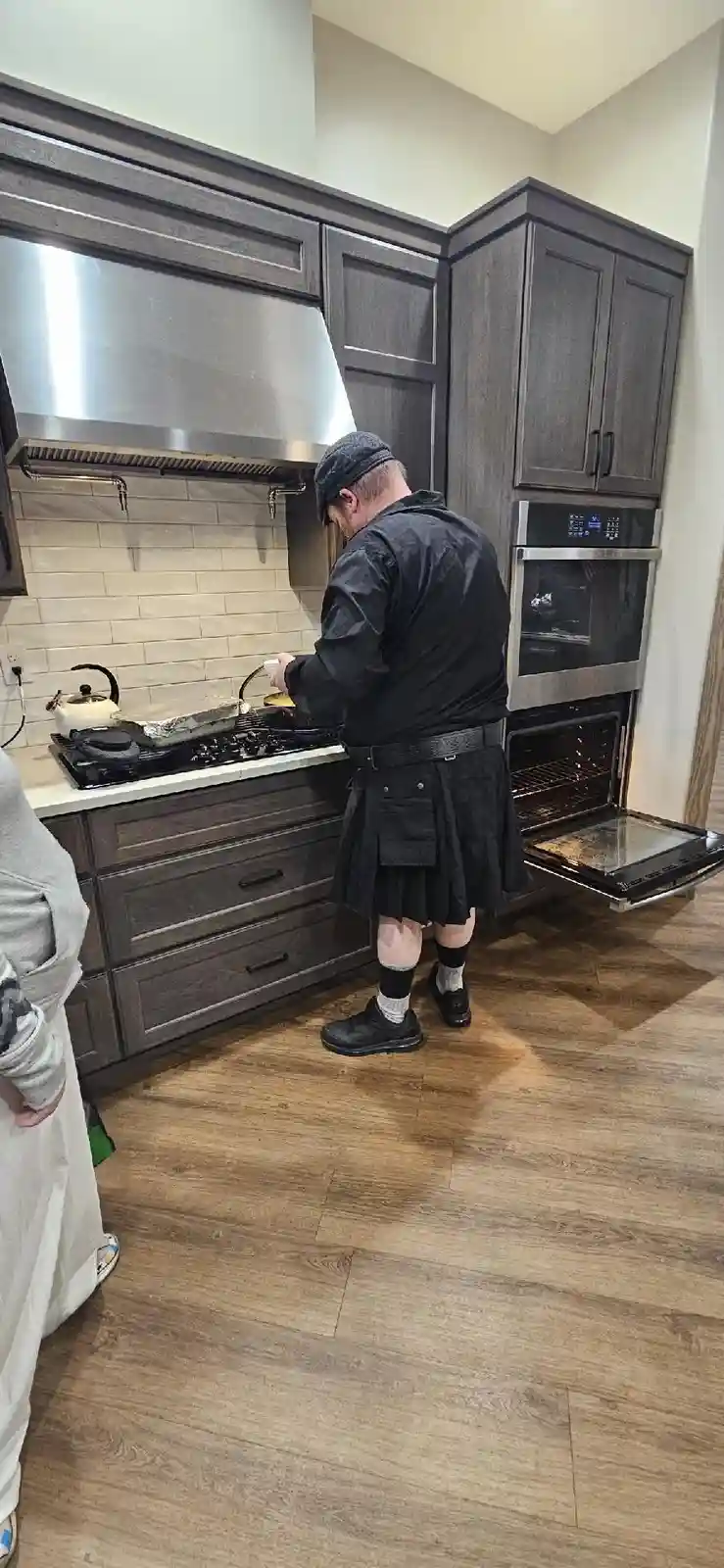 A man in a black kilt and cap is cooking at a modern kitchen stove while an oven door is open beside him. Another person is…