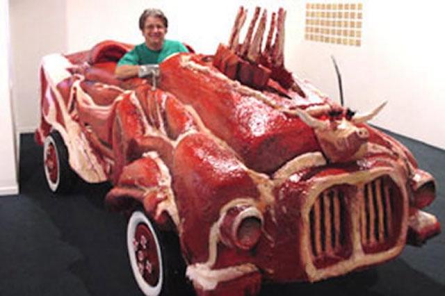 A man in a green shirt sits inside a car entirely sculpted from raw meat, with ribs and bull-like horns protruding from the…